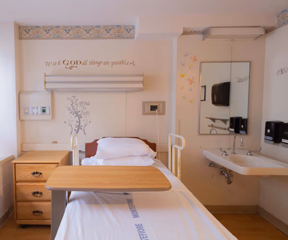 A hospital-style patient room with a single bed featuring white sheets and pillows, a wooden bedside table with three drawers, a wooden overbed table, a wall-mounted sink with a mirror above it, and soap dispensers on the wall. The walls have decorative text and designs, including the phrases 'With GOD all things are possible' and 'grow in faith,' along with colorful butterfly decals.