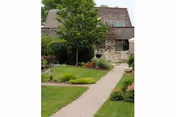 A paved walkway leading through a well-maintained garden with green grass, shrubs, and a tree, approaching a stone building with a brown roof.