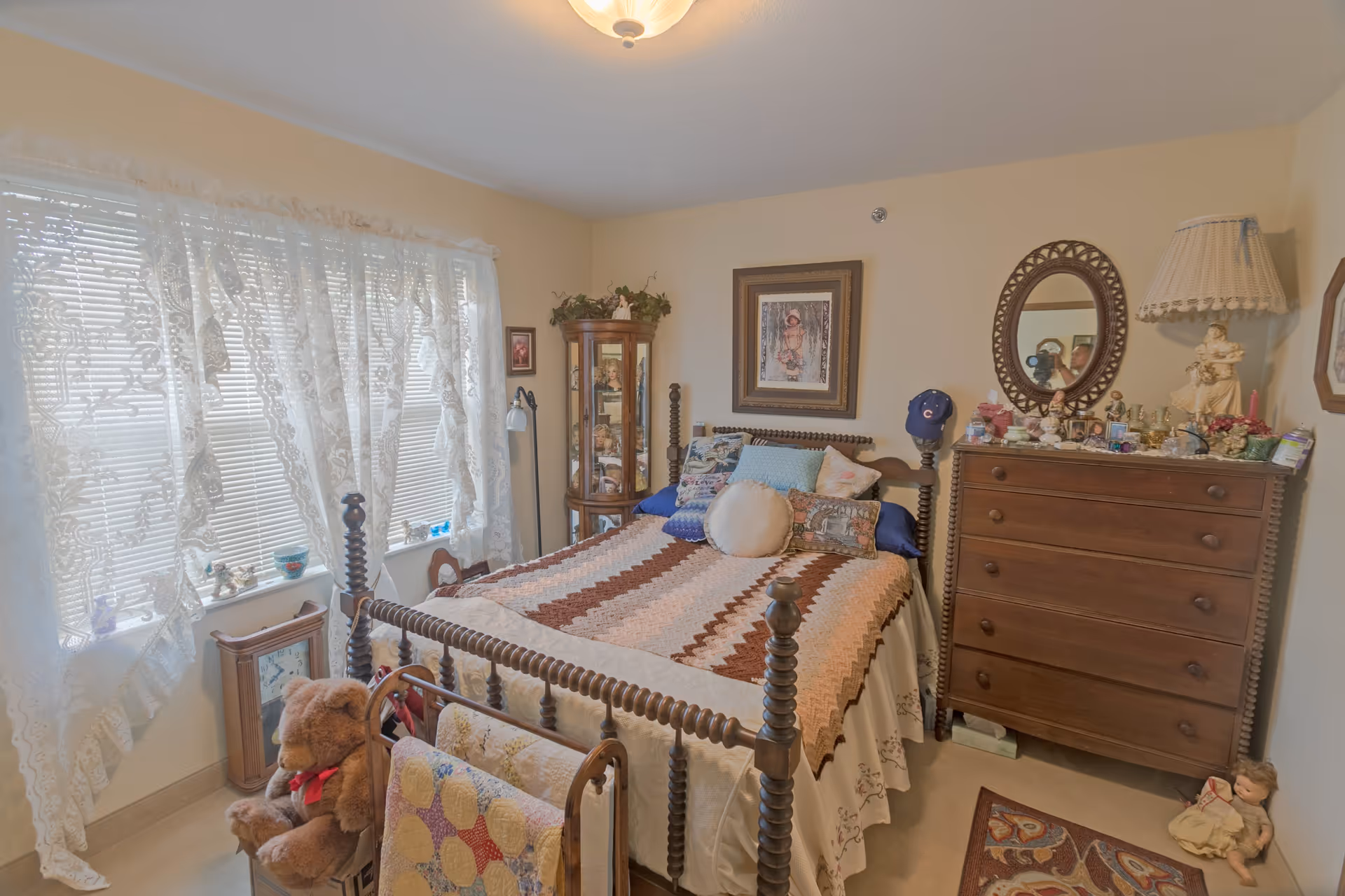 A cozy bedroom with a wooden bed covered by a crocheted blanket and multiple pillows. The room has lace curtains on the window, a wooden dresser with a mirror and various decorative items, a tall glass display cabinet, and a teddy bear sitting on a chair. The walls are adorned with framed pictures and the room has a warm, vintage feel.