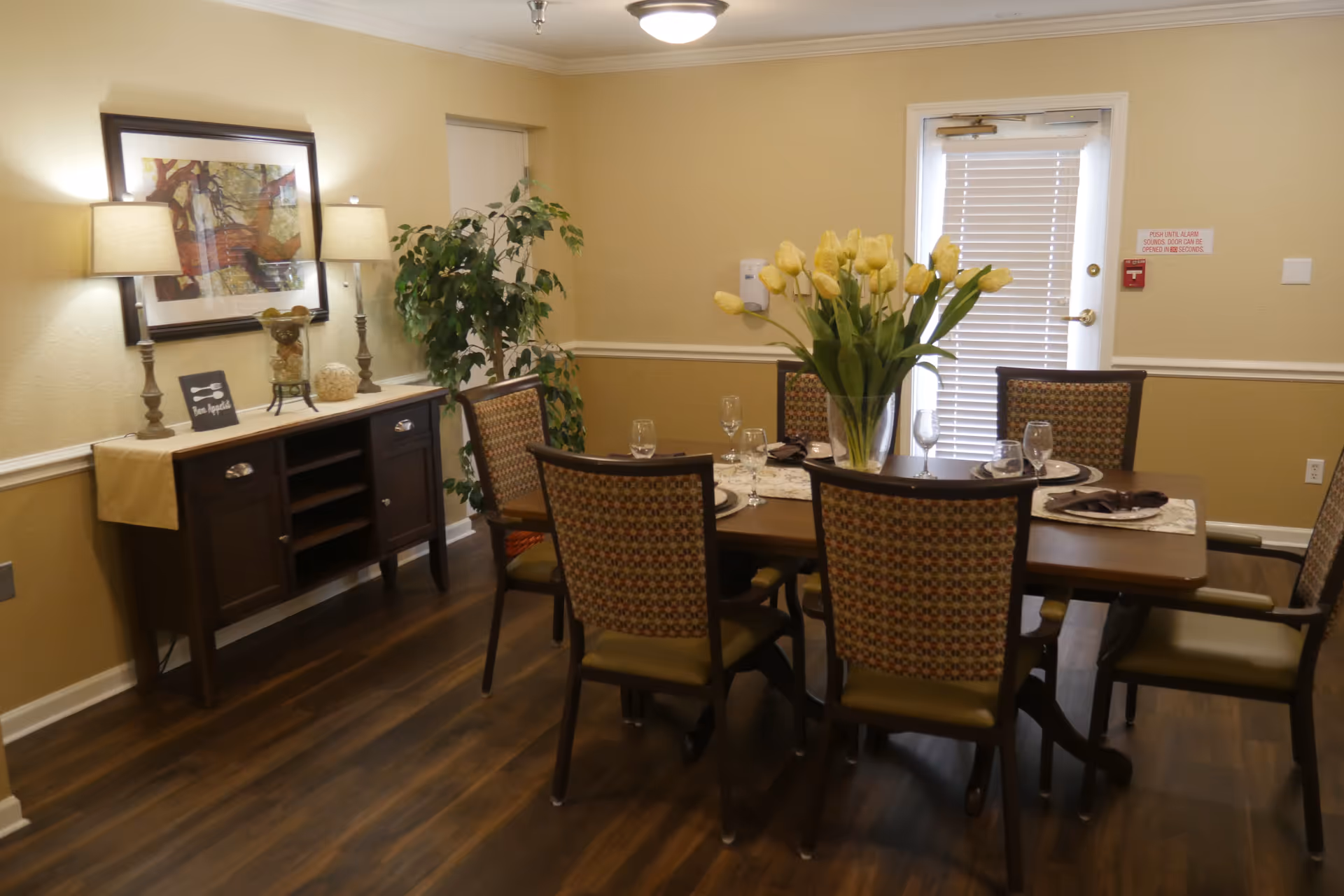 A dining room with a wooden table set for six people, featuring patterned chairs and a vase of yellow tulips as a centerpiece. There is a sideboard with two lamps, decorative items, and a framed artwork on the wall. A potted plant is in the corner, and a door with blinds is visible in the background.
