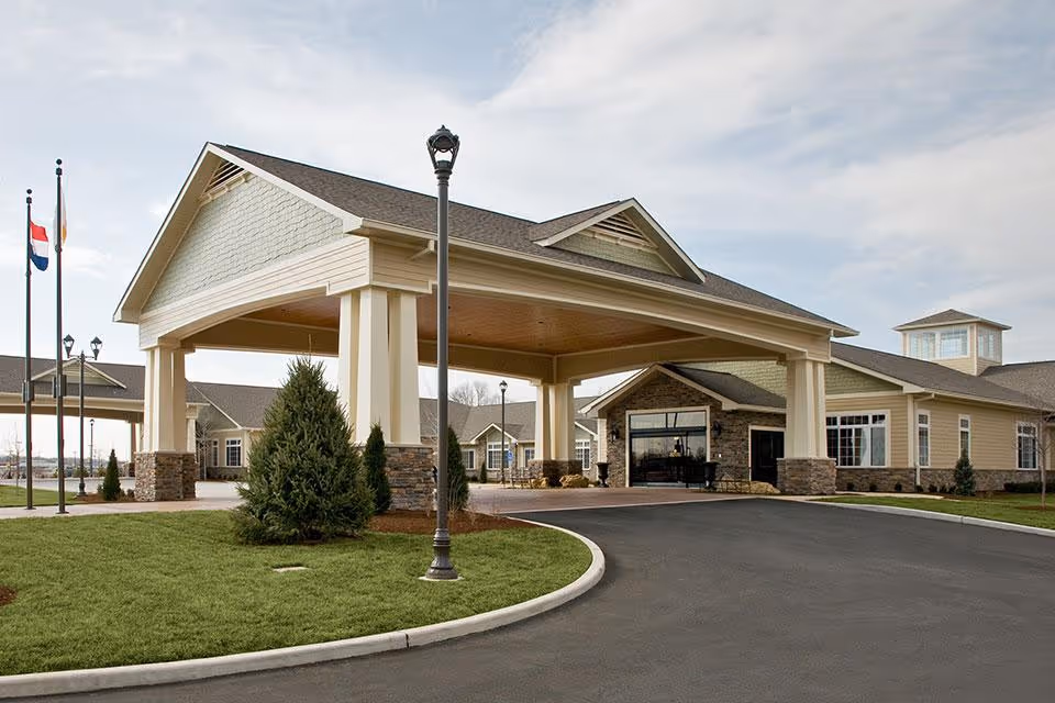 Exterior view of the entrance to Life Care Center of Cape Girardeau, featuring a large covered drop-off area with stone and beige pillars, a curved driveway, green lawn with small trees, and flagpoles with flags.