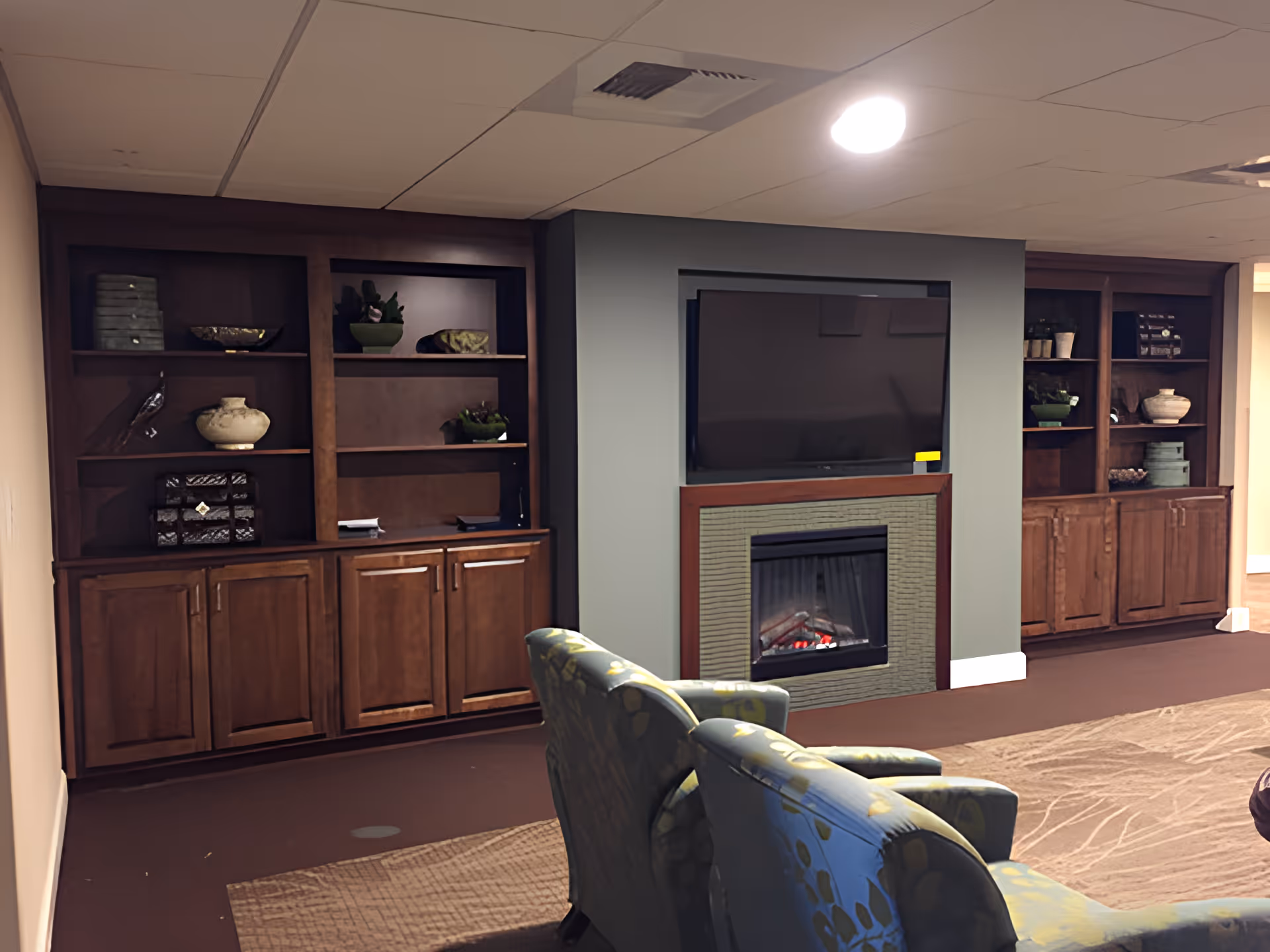 A cozy living room area featuring a wall-mounted flat screen TV above a modern electric fireplace, flanked by two wooden built-in shelving units with decorative items. In the foreground, there are two upholstered armchairs with a floral pattern, positioned on a carpeted floor.