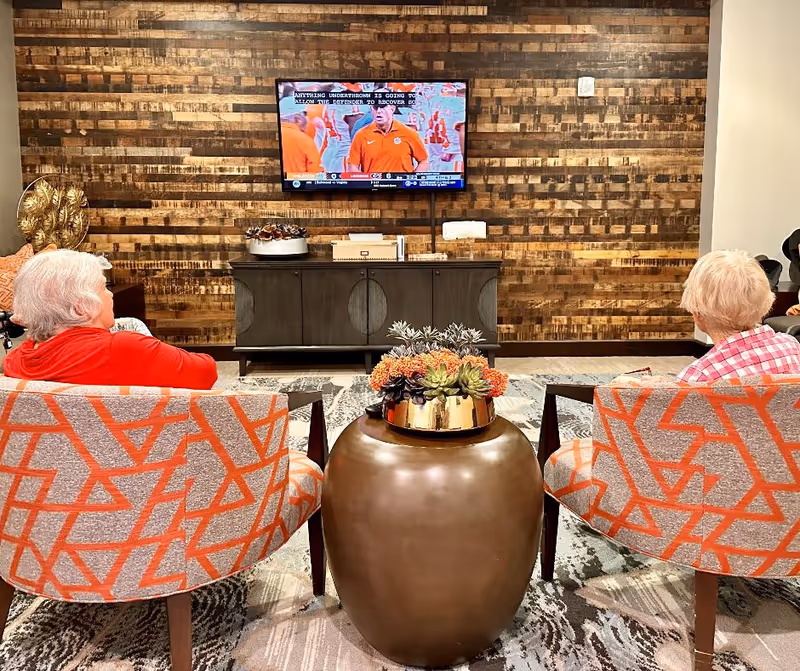 Two elderly women sitting in orange and gray patterned chairs watching a football game on a wall-mounted TV in a cozy room with a wooden accent wall and a round bronze table with a floral arrangement between them.