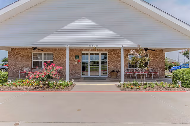 Front exterior view of a single-story brick building with a white peaked roof and a covered entrance. There are two sets of glass double doors in the center, flanked by windows. Flower beds with blooming pink flowers and shrubs line the walkway leading to the entrance. Several metal chairs are placed on either side of the entrance under the covered area. Cars are visible parked on the right side in the background.