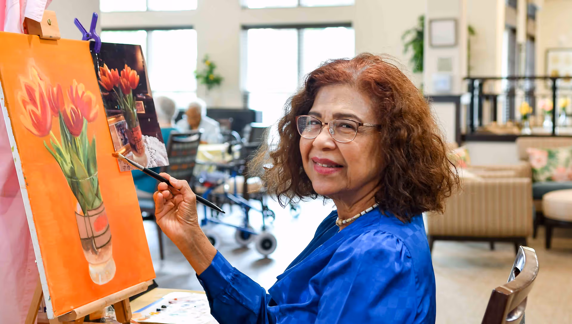 An elderly woman with glasses and curly hair is painting a picture of red tulips in a vase on an easel inside a well-lit room with large windows and comfortable seating in the background.