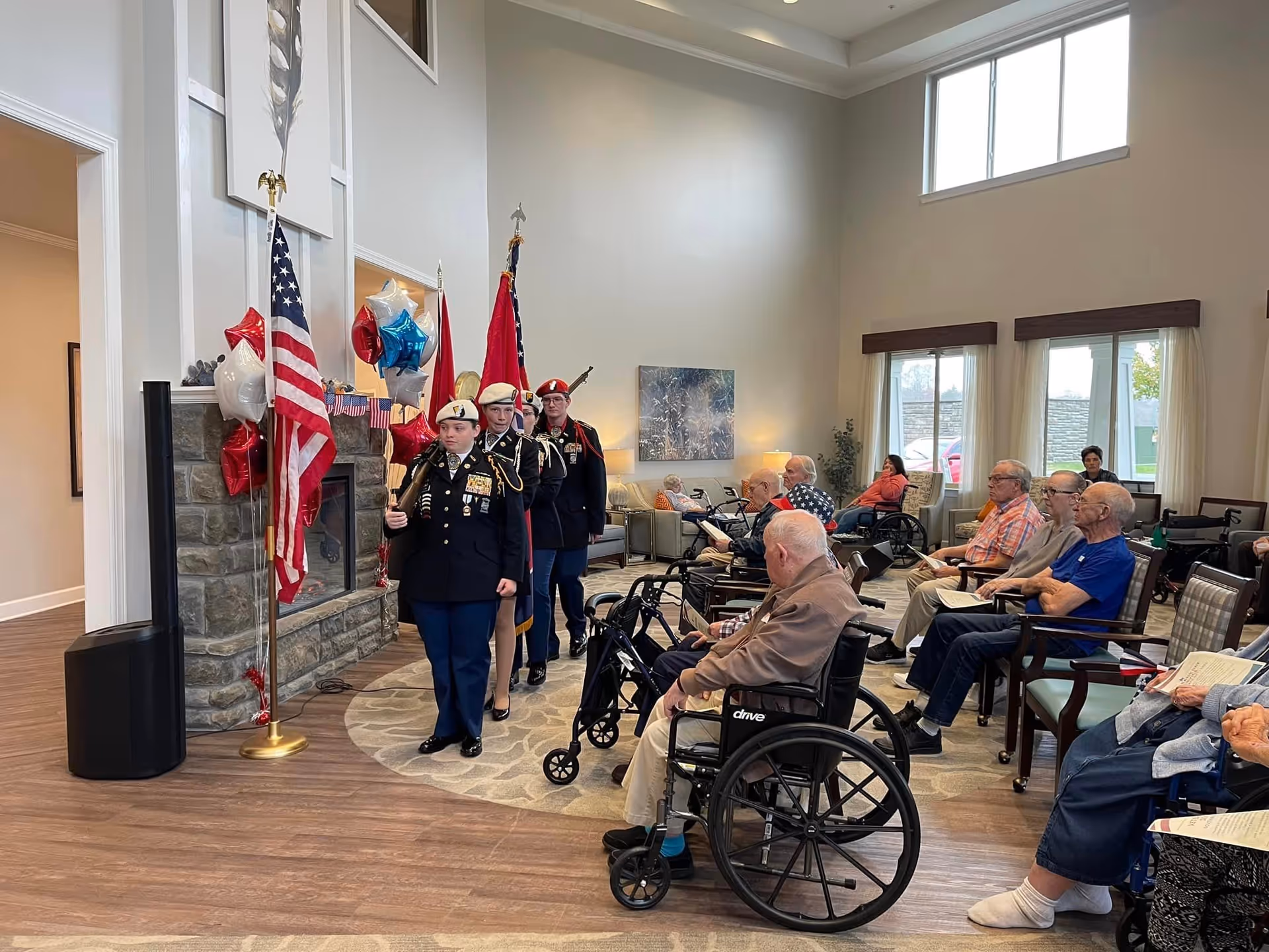 A group of uniformed individuals carrying flags march in a senior living facility common area where elderly residents, some in wheelchairs, are seated and watching the procession. The room has large windows, a stone fireplace decorated with red, white, and blue balloons, and comfortable seating.