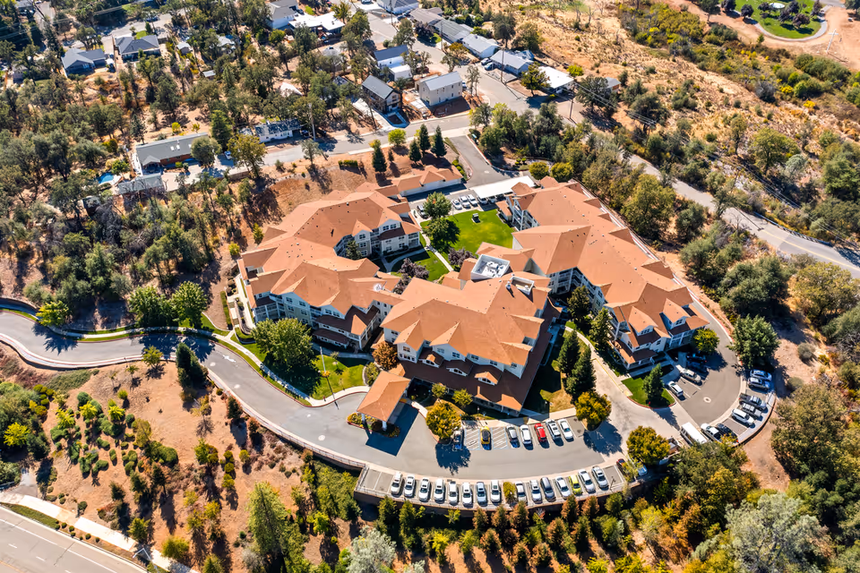 Aerial view of a large senior living facility with multiple connected buildings featuring red-tiled roofs, surrounded by trees and greenery. There is a parking lot with numerous cars in front of the buildings and a curved driveway leading to the entrance. Residential houses and roads are visible in the background.
