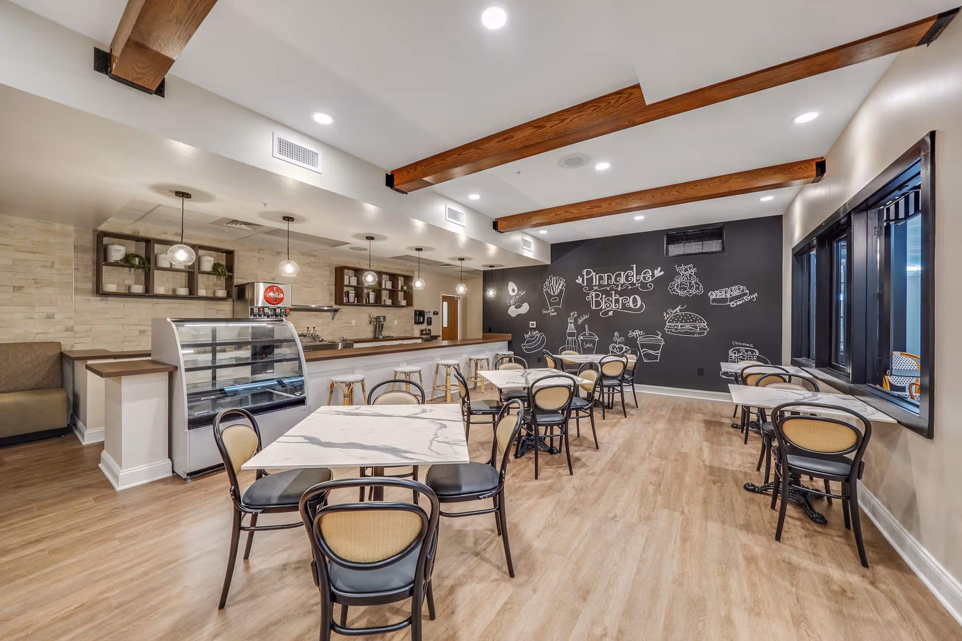 Interior view of a dining area named Pinnacle Bistro with several tables and chairs, a counter with bar stools, pendant lights, and a blackboard wall featuring food illustrations and the text 'Pinnacle Bistro'.