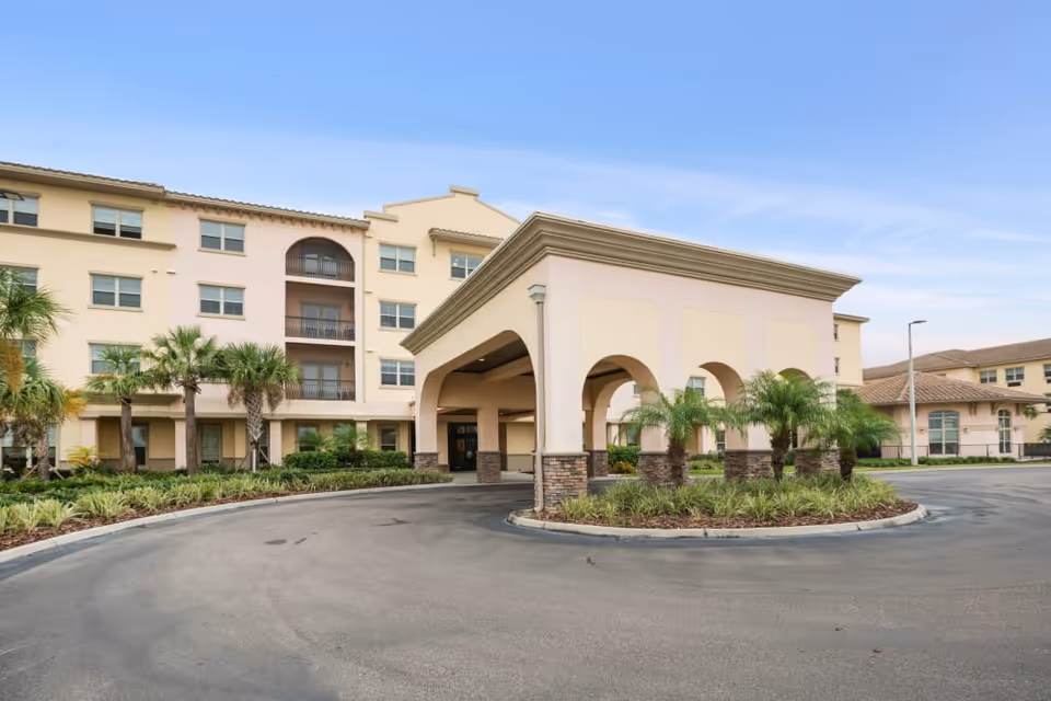 Front exterior of a multi-story senior living building with a covered porte-cochere, palm trees, and a circular driveway.