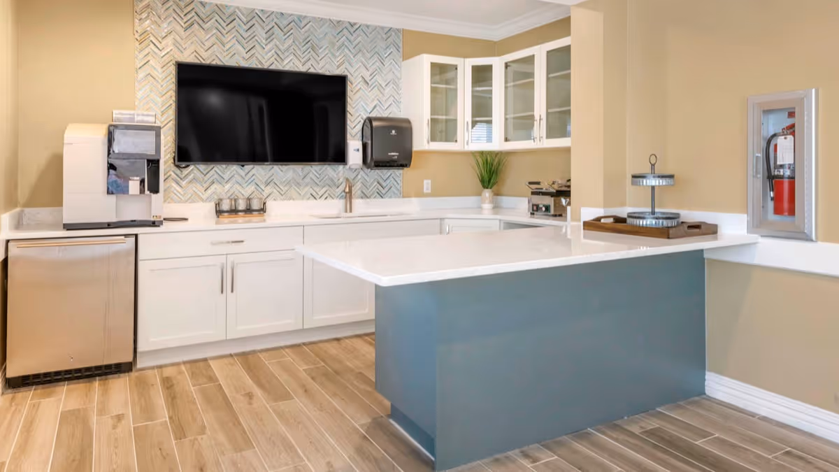 A modern kitchen area with white cabinets, a large flat-screen TV mounted on a patterned backsplash, a coffee machine, a paper towel dispenser, a small sink, and a kitchen island with a white countertop. The floor is covered with wood-look tiles, and there is a fire extinguisher mounted on the wall.