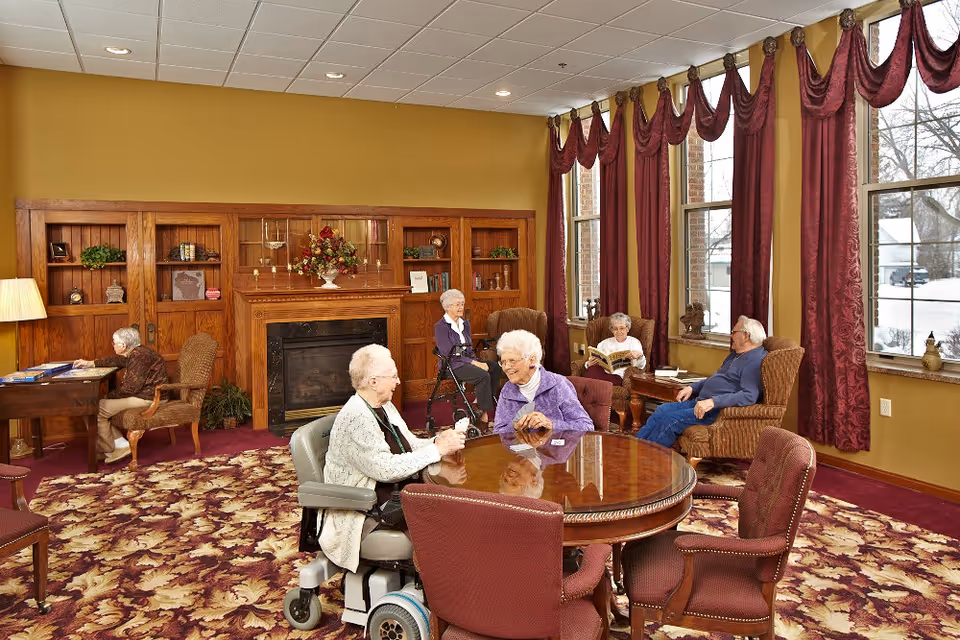 Several elderly residents socialize and read in a carpeted senior living common room with a fireplace, round table, and large windows with burgundy drapes.