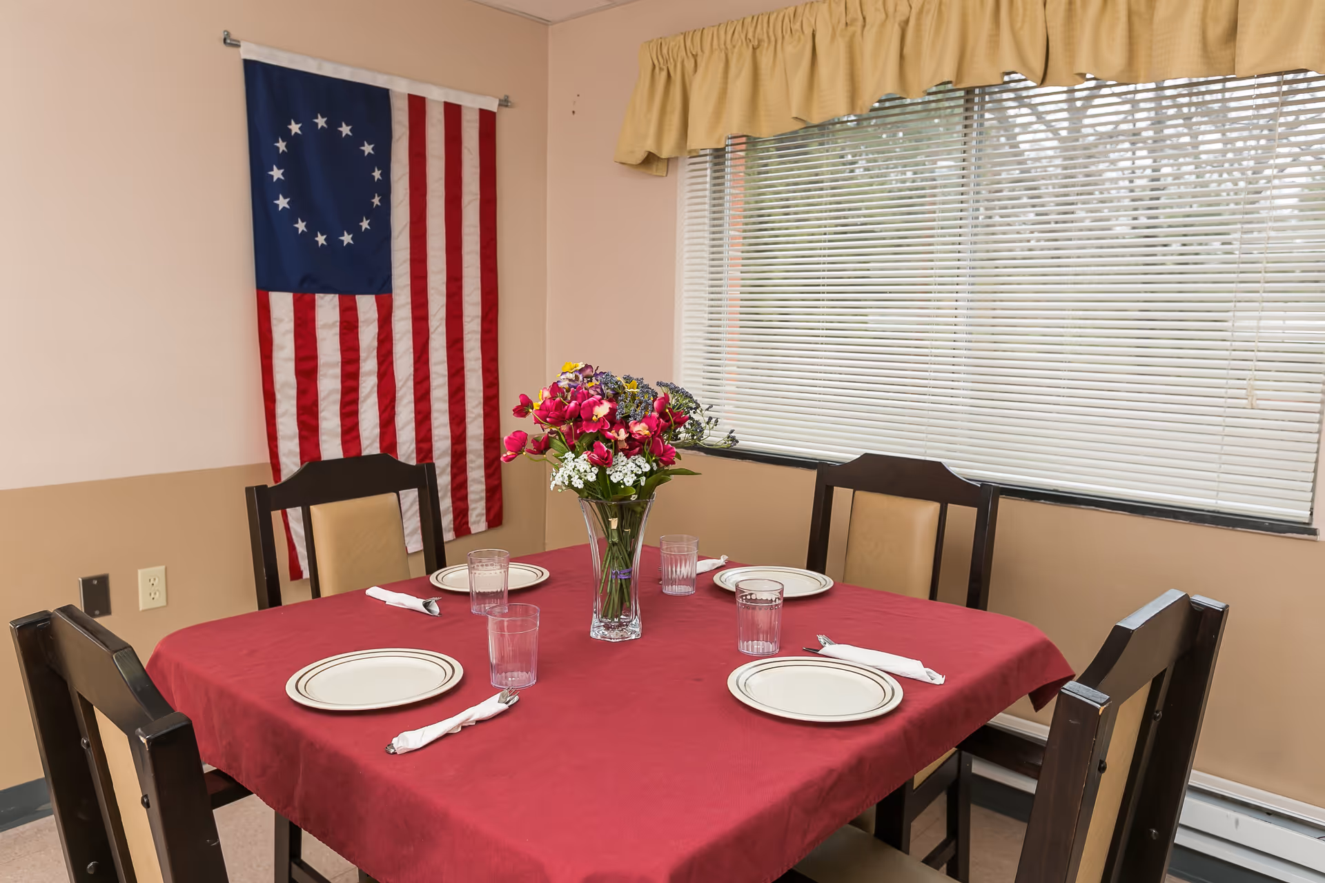 A dining table set for four with a red tablecloth, four plates, four glasses, and four napkins. A vase with colorful flowers is placed in the center of the table. Behind the table, there is a window with closed blinds and a yellow valance. On the wall to the left, a Betsy Ross American flag is hanging.