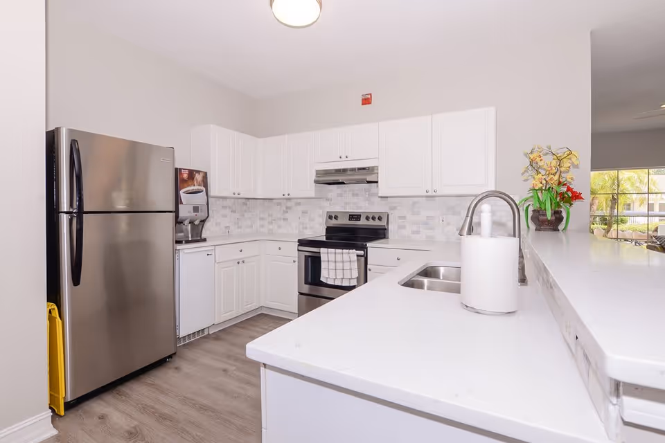 A clean, modern kitchen with white cabinets, a stainless steel refrigerator, stove, and dishwasher. There is a white countertop with a sink and a paper towel holder. A coffee machine is placed on the counter near the refrigerator. A vase with yellow and red flowers is on the counter near a window showing an outdoor view with palm trees.