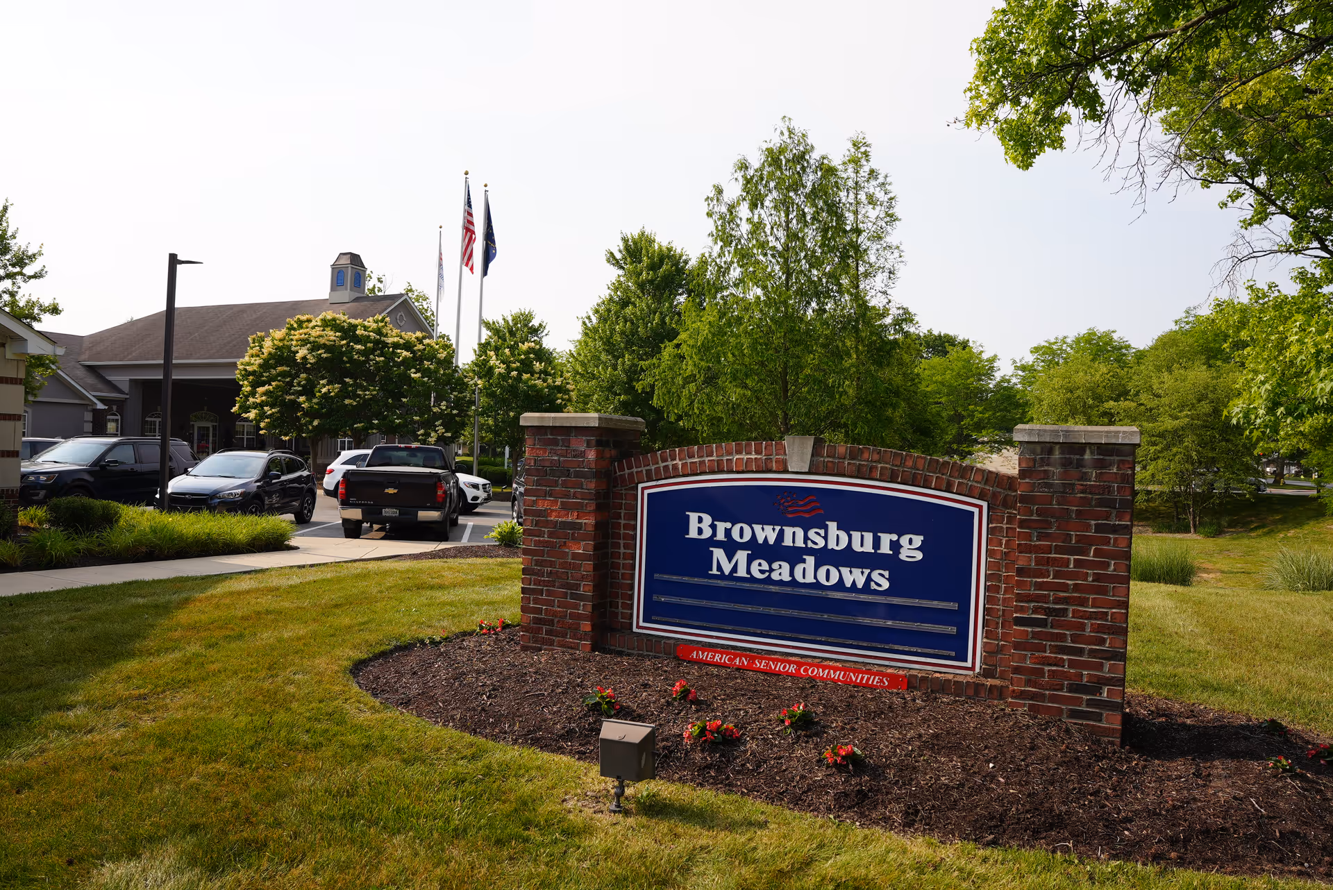 Outdoor view of Brownsburg Meadows senior living facility sign made of brick with blue and white text, surrounded by grass, flowers, trees, and parked cars in the background.