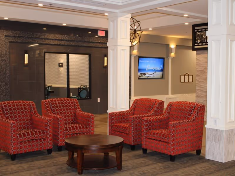 A cozy seating area in a senior living facility with four red patterned armchairs arranged around a round wooden coffee table. The room features white pillars, a wall-mounted TV displaying a summer message, and soft lighting fixtures on the walls.