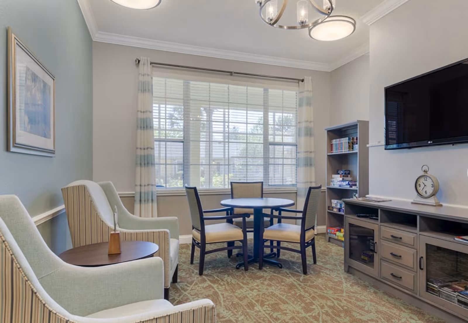 A cozy sitting area in a senior living facility with two upholstered armchairs around a small round wooden table, a round table with four chairs near a large window with striped curtains, a bookshelf with board games and books, a wall-mounted flat screen TV, and a cabinet with drawers and shelves. The room has soft lighting and a carpeted floor.