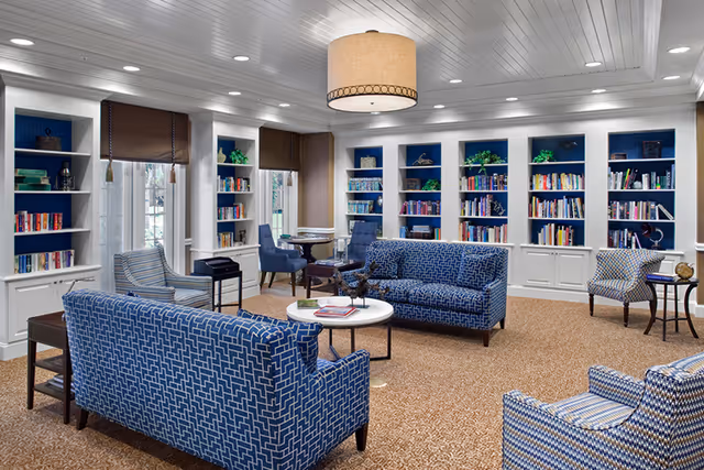 A cozy and well-lit library or reading room with built-in white bookshelves filled with books and decorative items. The room features blue patterned sofas and armchairs arranged around a small round white coffee table. There are windows with brown roman shades, a beige carpet, and a large ceiling light fixture.