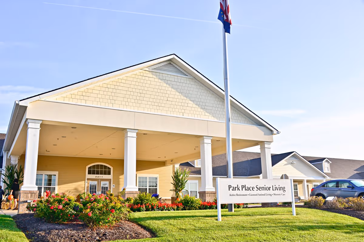 Exterior view of Park Place Senior Living facility with a covered entrance supported by white columns, a well-maintained garden with flowers in front, a flagpole, and a sign displaying the facility's name and services. A car is parked on the right side.