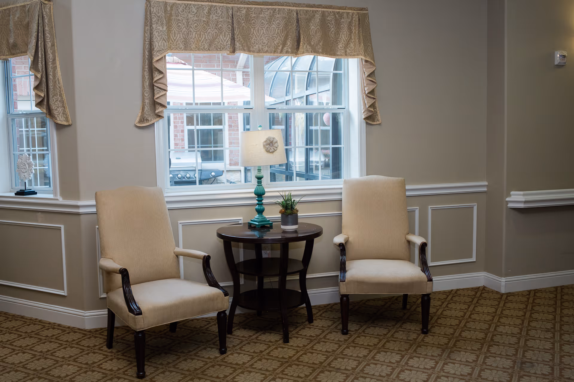 Two beige upholstered armchairs with dark wooden legs and armrests are placed on either side of a small round wooden table. The table holds a turquoise lamp with a beige lampshade and a small potted plant. Behind the chairs is a large window with beige patterned valances, showing an outdoor view of a brick building and a covered patio area.