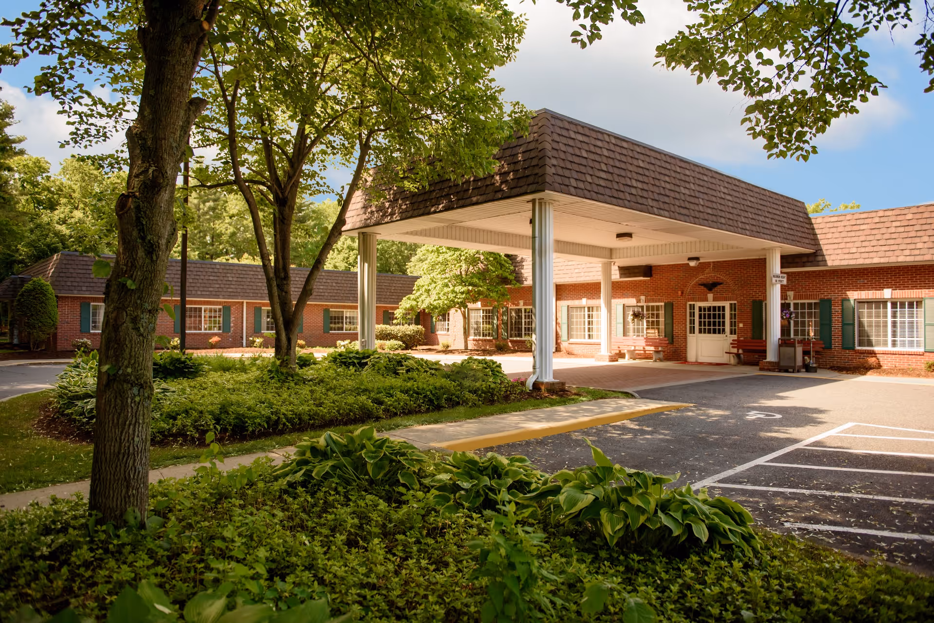 Exterior view of a single-story brick building with a covered entrance supported by white columns. The building is surrounded by green trees and shrubs, with a paved driveway and parking spaces in front. The sky is partly cloudy with blue patches visible.