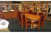 A cozy library or reading room with wooden bookshelves filled with books lining the walls. In the center, there is a wooden table surrounded by six cushioned chairs with red upholstery. The room has a carpeted floor and framed pictures on the walls.