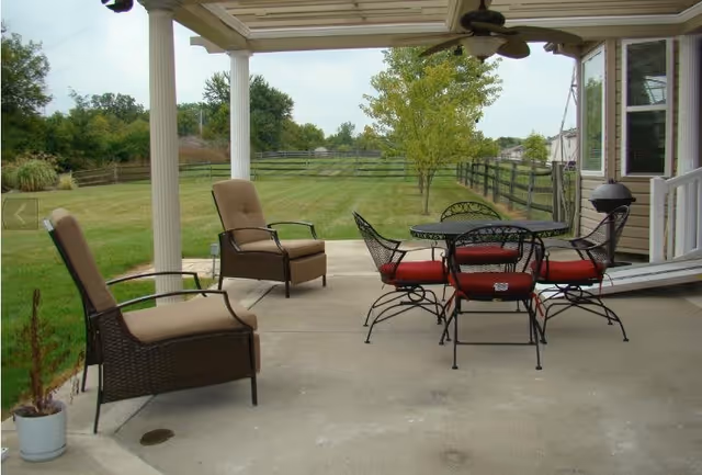 Covered patio with outdoor seating and a round table overlooking a fenced grassy yard.