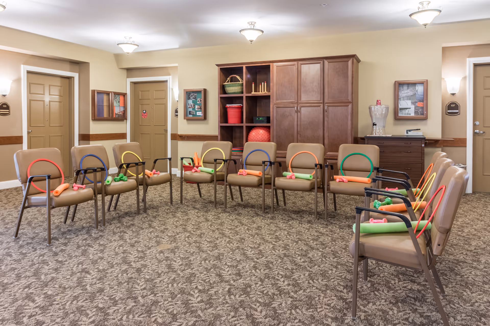 A room with a circle of brown chairs arranged for group activity, each chair holding colorful exercise equipment including small dumbbells, foam noodles, and hoops. The room has beige walls, carpeted floor, wooden cabinets, and three closed doors. There are framed pictures and wall sconces on the walls, and a water dispenser on a cabinet.