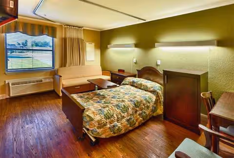 A furnished senior living bedroom with a bed, sofa, desk, and window.