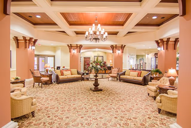 A spacious and elegant living room area with a patterned carpet, multiple sofas and armchairs arranged around a central round table with a floral arrangement. The room features warm lighting, decorative columns, and a chandelier hanging from a coffered ceiling with wooden accents.