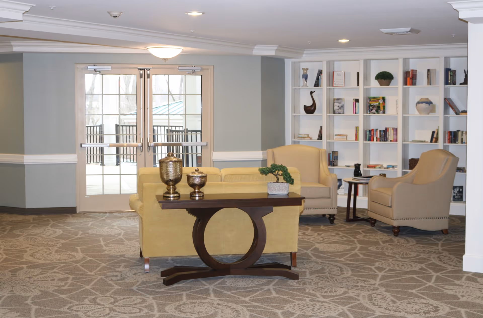 A cozy senior living common area with a yellow sofa, two beige armchairs, a dark wooden table with decorative items, and a white built-in bookshelf filled with books and ornaments. Double glass doors lead to an outdoor area.