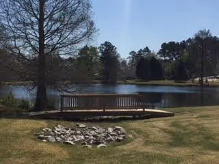 A peaceful outdoor scene featuring a small wooden dock extending over a calm pond surrounded by grass and trees under a clear sky.