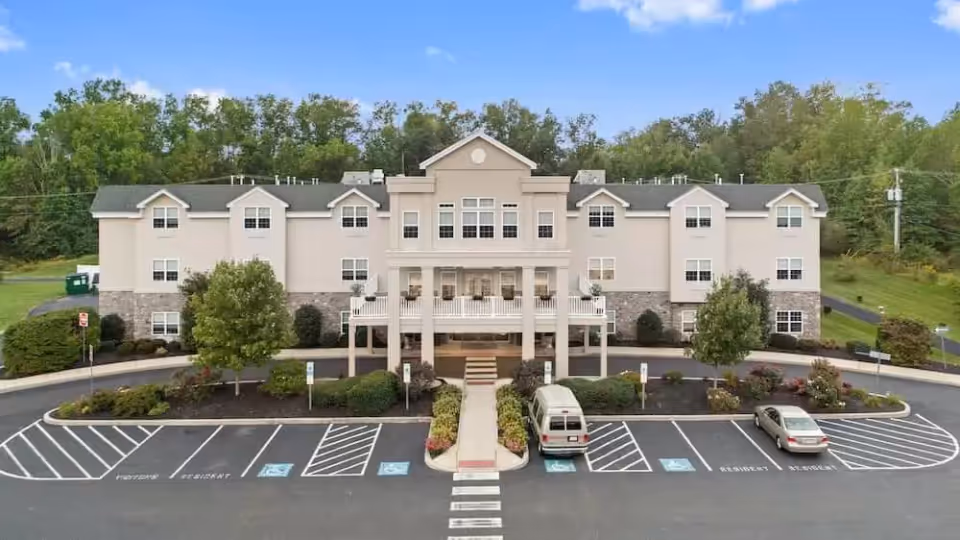 Front exterior view of a three-story senior living facility named Hidden Meadows On the Ridge, featuring a central entrance with a covered porch, surrounded by landscaped greenery and a parking lot with designated parking spaces including handicapped spots.