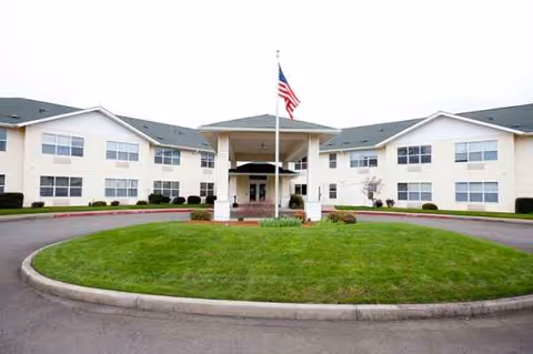 Two-story beige senior living building with central entrance, flagpole, and a circular grassy driveway island.