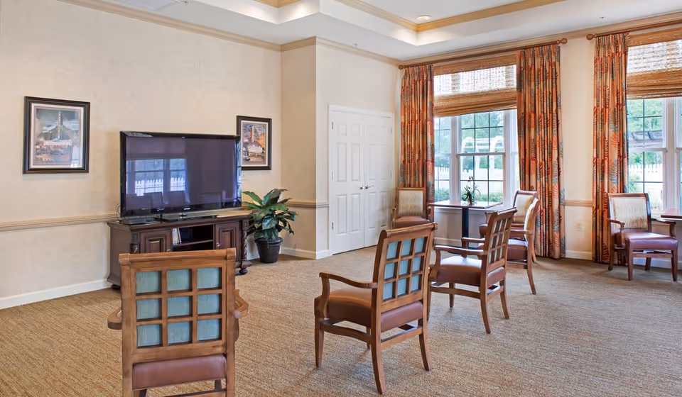 A bright and spacious common area with several wooden chairs arranged facing a large flat-screen TV on a wooden stand. The room features beige walls, carpeted flooring, two framed pictures on the wall, a potted plant, and large windows with patterned curtains allowing natural light to enter.