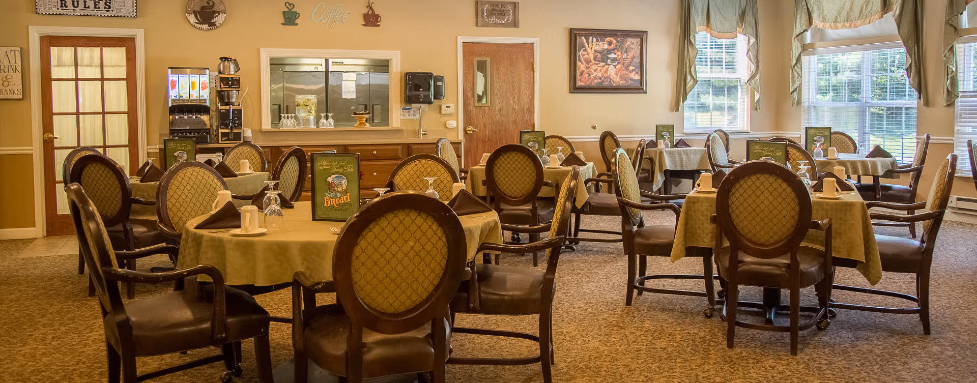 A dining room in a senior living facility with multiple round tables covered with beige tablecloths, each surrounded by cushioned chairs. The tables are set with napkins, glasses, and small menu cards. Large windows with curtains allow natural light to fill the room. The walls are decorated with framed pictures and signs, and there is a beverage station with a soda dispenser and cups in the background.
