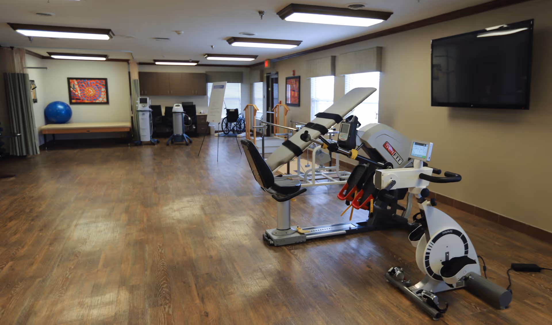 A spacious rehabilitation room with wooden flooring featuring exercise equipment including a stationary bike and a leg press machine. The room has several windows with blinds, a large flat-screen TV mounted on the wall, a blue exercise ball, a bench, and cabinets along the back wall. There is also a wheelchair and a whiteboard in the background.