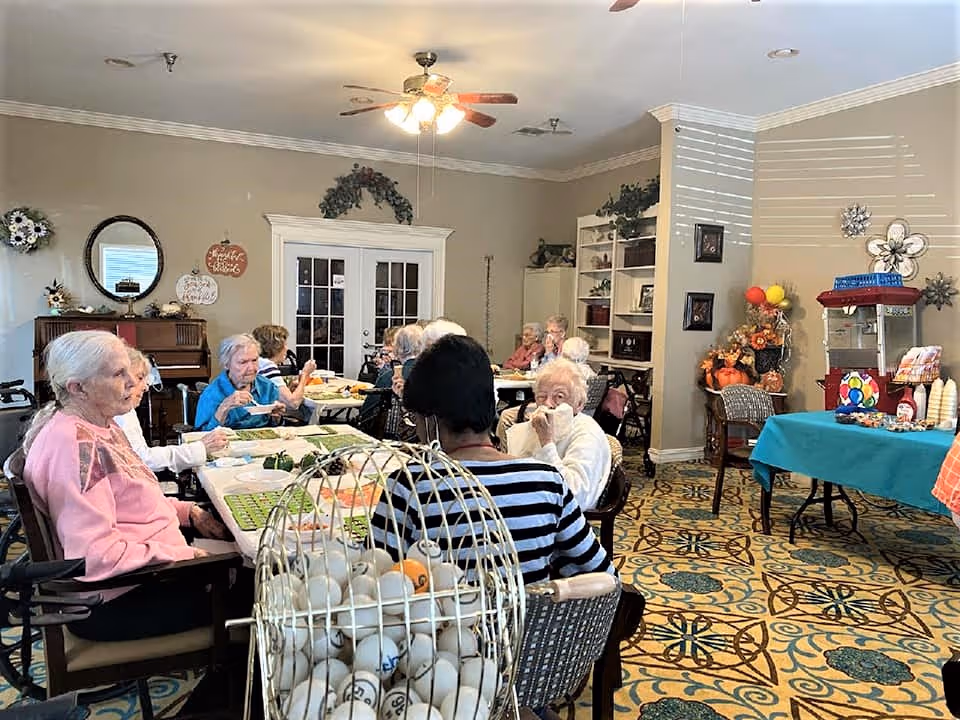 A group of elderly people sitting around tables in a well-lit room playing bingo. The room has patterned carpet, ceiling fans, and decorative items on the walls and shelves. There is a bingo cage with balls in the foreground and a table with snacks and a popcorn machine on the right side.
