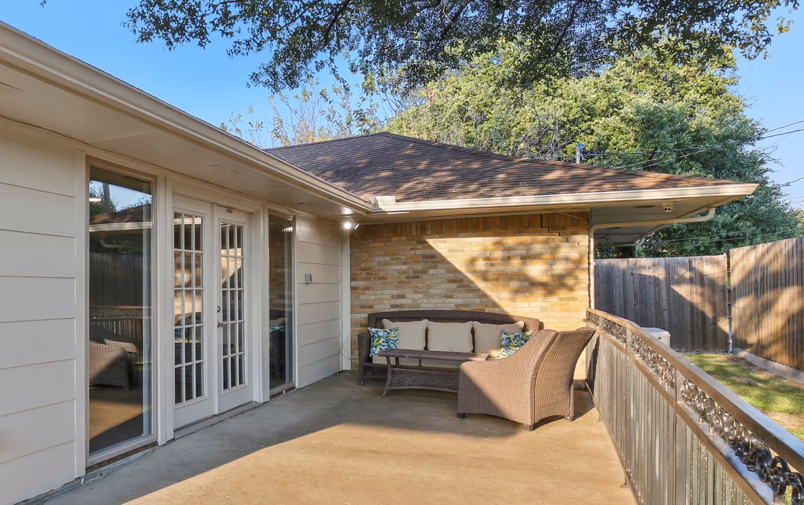 Outdoor patio area with a wicker sofa and chair set with cushions, adjacent to a building with beige siding and glass French doors, surrounded by a wooden fence and trees in the background.