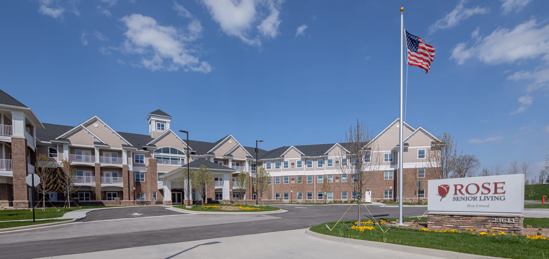 Exterior view of Rose Senior Living Beachwood facility on a clear day with blue sky. The building is three stories tall with a combination of brick and beige siding, multiple windows, and balconies. There is a driveway leading to the entrance, a flagpole with an American flag, and a sign displaying the facility's name.