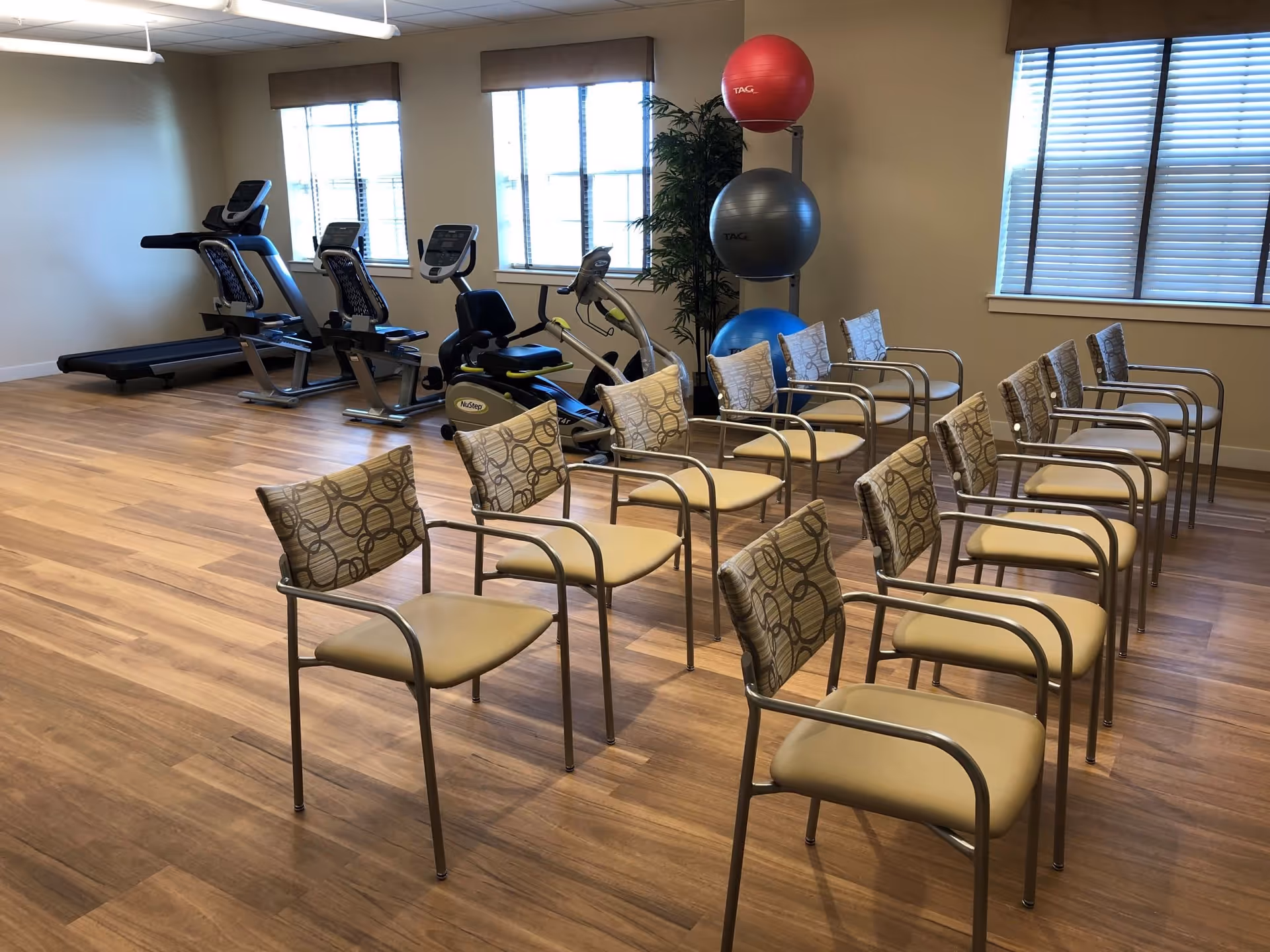 A fitness room with exercise equipment including treadmills and stationary bikes, several chairs arranged in rows, and a rack holding three large exercise balls in red, gray, and blue. The room has wooden flooring, beige walls, and windows with blinds.