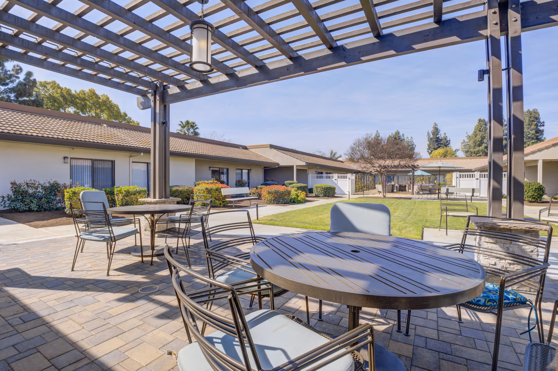 Outdoor patio area at Park Visalia Assisted Living & Memory Care with round metal tables and chairs under a pergola. The patio overlooks a grassy courtyard surrounded by single-story buildings with tiled roofs and landscaped bushes.