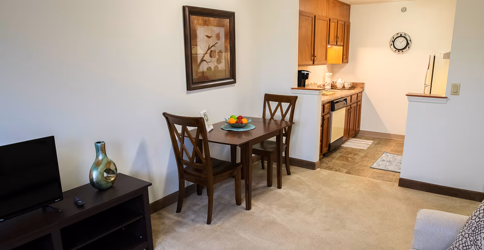 A small dining area with a wooden table and two chairs, a bowl of fruit on the table, a framed picture on the wall, and a view into a kitchen with wooden cabinets, a dishwasher, and a clock on the wall. A TV and decorative vase are on a stand to the left, and part of a sofa is visible on the right.