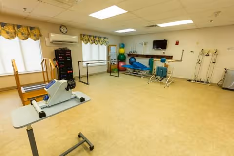 A spacious physical therapy room with exercise equipment including a recumbent bike, parallel bars, therapy balls, a treatment table, and a wall-mounted TV. The room has large windows with yellow patterned valances and bright overhead lighting.