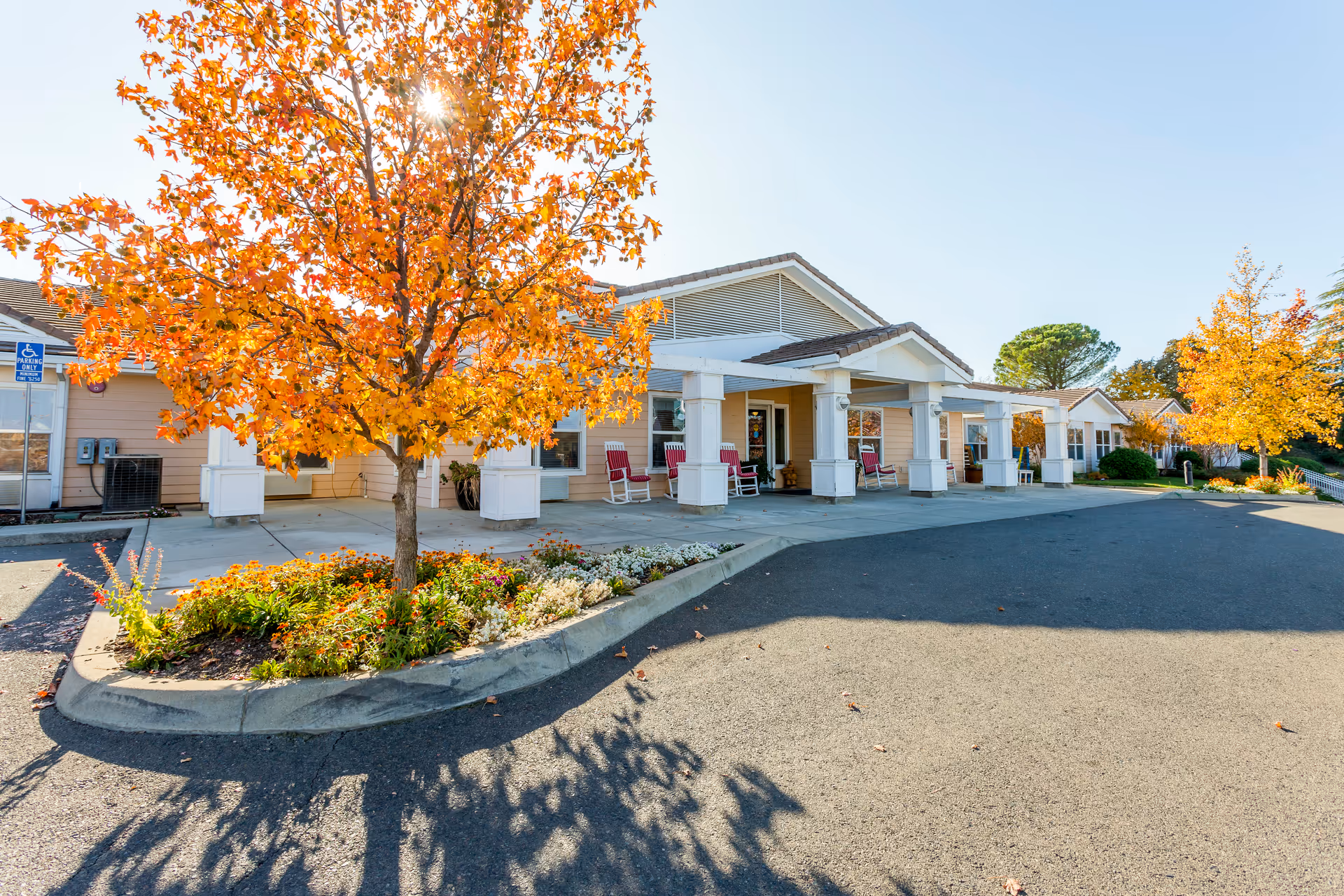 Front entrance of a senior living building with a covered porch, rocking chairs, autumn trees, and a curved driveway.