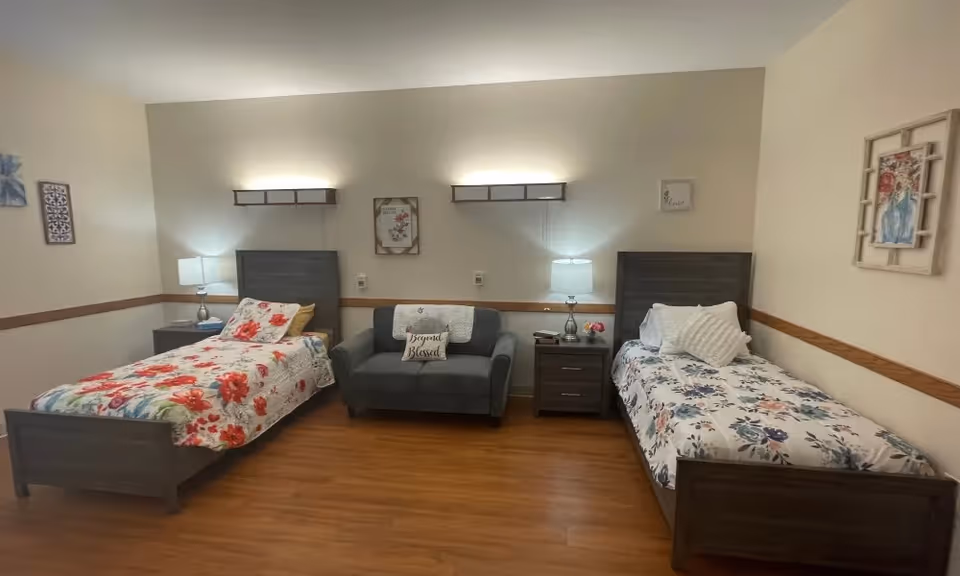 A senior living facility bedroom with two single beds, each with floral bedding. Between the beds is a small gray loveseat with a decorative pillow that says 'Beyond Blessed.' There are two nightstands with lamps on either side of the loveseat. The walls are decorated with framed floral artwork and the floor is wooden.