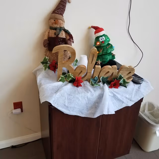 A wooden cabinet covered with a white cloth, decorated with a wooden sign spelling 'Believe' surrounded by artificial green leaves and red flowers. Behind the sign, there is a small plush Christmas tree wearing a Santa hat with lights and a snowman figure holding a book. A remote control and a white object are also on the cabinet. The background shows a beige wall with a visible electrical outlet and a trash bin to the right.