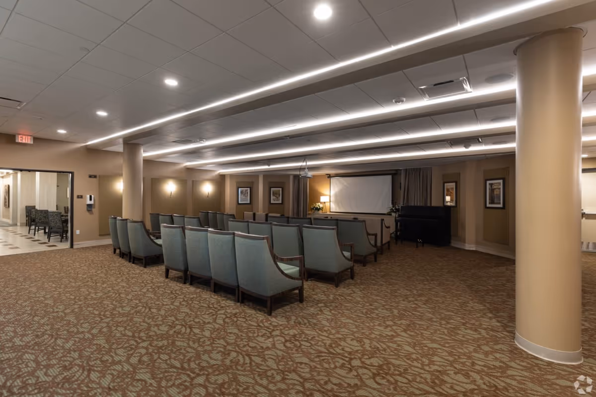A carpeted community activity room with rows of upholstered chairs facing a projector screen and a piano.