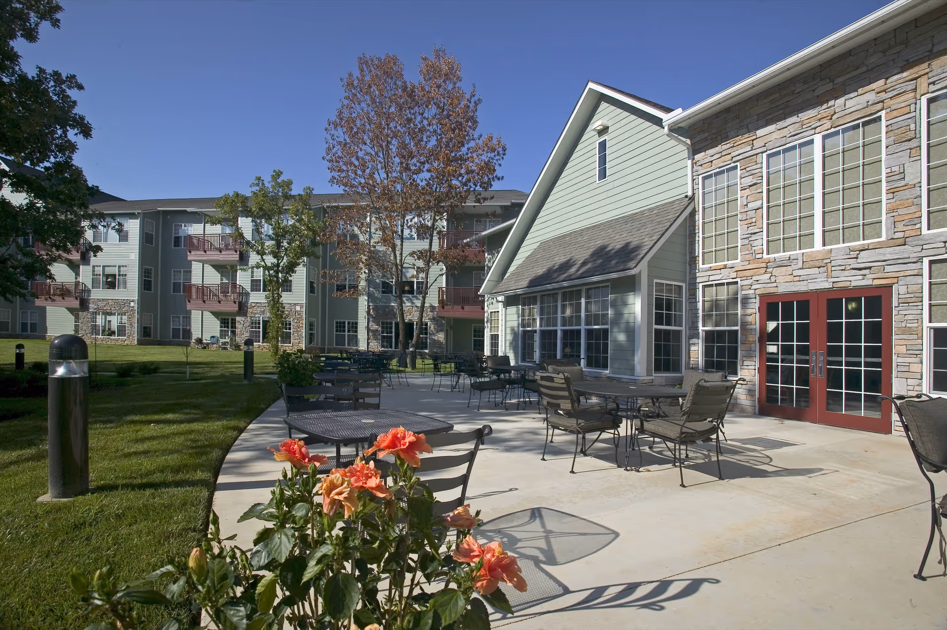 Outdoor patio area at Plaza At Highlands Crossing with metal tables and chairs on a concrete surface, surrounded by green grass and trees. The building exterior features stone and light green siding with large windows and red double doors under a clear blue sky.