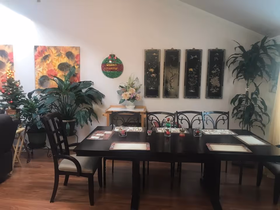 Dining room with a dark wooden table set with placemats and chairs, decorative wall art and several potted plants.
