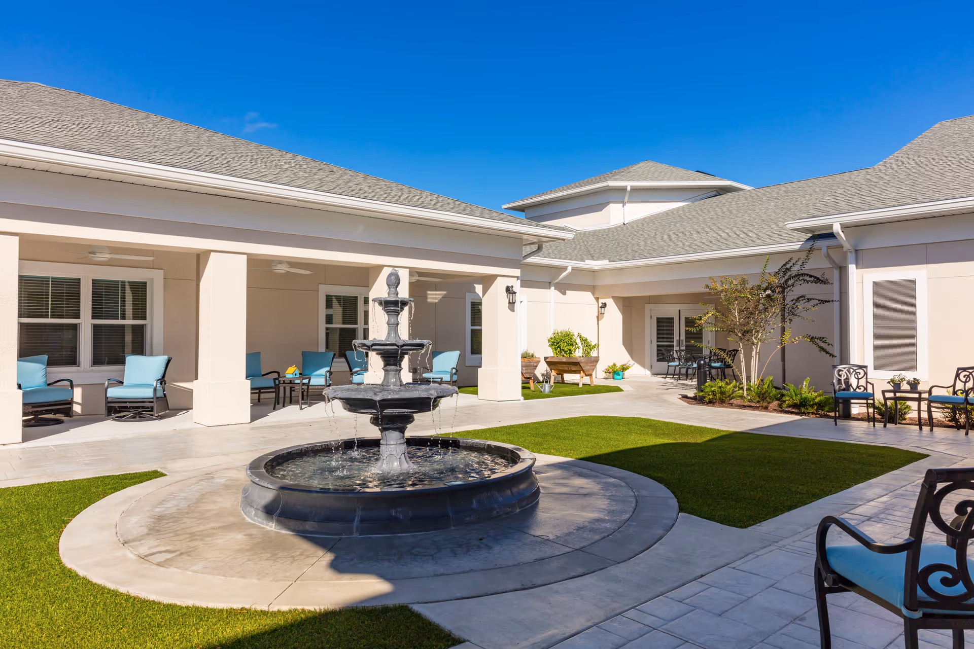 Outdoor courtyard area of a senior living facility with a central three-tiered water fountain, surrounded by a circular paved walkway and green grass patches. The courtyard is enclosed by a single-story building with beige walls and a gray shingled roof. There are several seating areas with blue cushioned chairs and small tables placed under covered walkways and near the building walls. The sky is clear and blue.